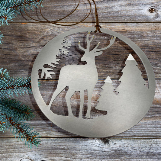Stainless steel ornament depicting a deer in the woods. The antlers of the deer and the tree tops are covered in white and the antlers have golden tips